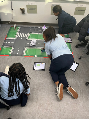 Three girls sit on the floor around a mat showing roads in a town as they watch their Photon robot move around.