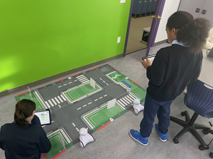 One girl stands and another sits as their Photon drives around the mat with their town.
