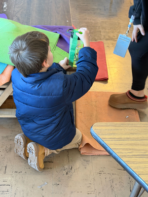 A boy in a blue winter coat sits on his heels as he measures something with his green folded fraction strip.