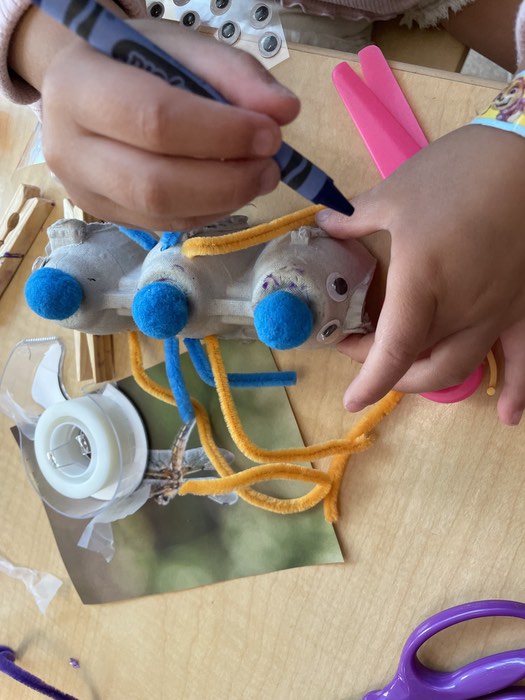 A child's hands hold a piece of egg carton 3 lumps long that they have turned into a caterpillar by attaching blue tufts to each hump, chenille (pipe cleaner) legs, and google eyes. They are holding a blue crayon.