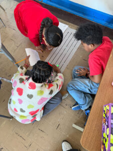 Three kids lean into a large paper with 5 blank number lines on the floor. They are watching an Ozobot go down one of the lines.