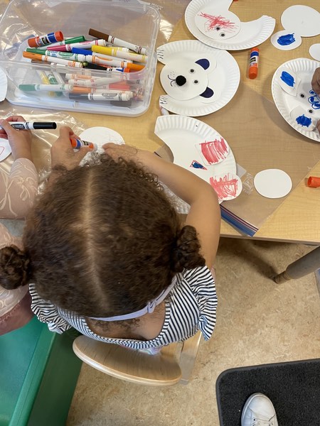 A little girl is holding a red marker as she starts to color in a polar bear she is making from a paper plate.
