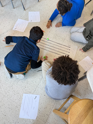 Looking over the shoulders of two boys, we see a large piece of paper on the floor with several number lines on it. An Ozobot travels along one line as on of the children leans in closer.