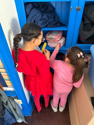 Two girls use a paper strip to measure something on a blue shelf.
